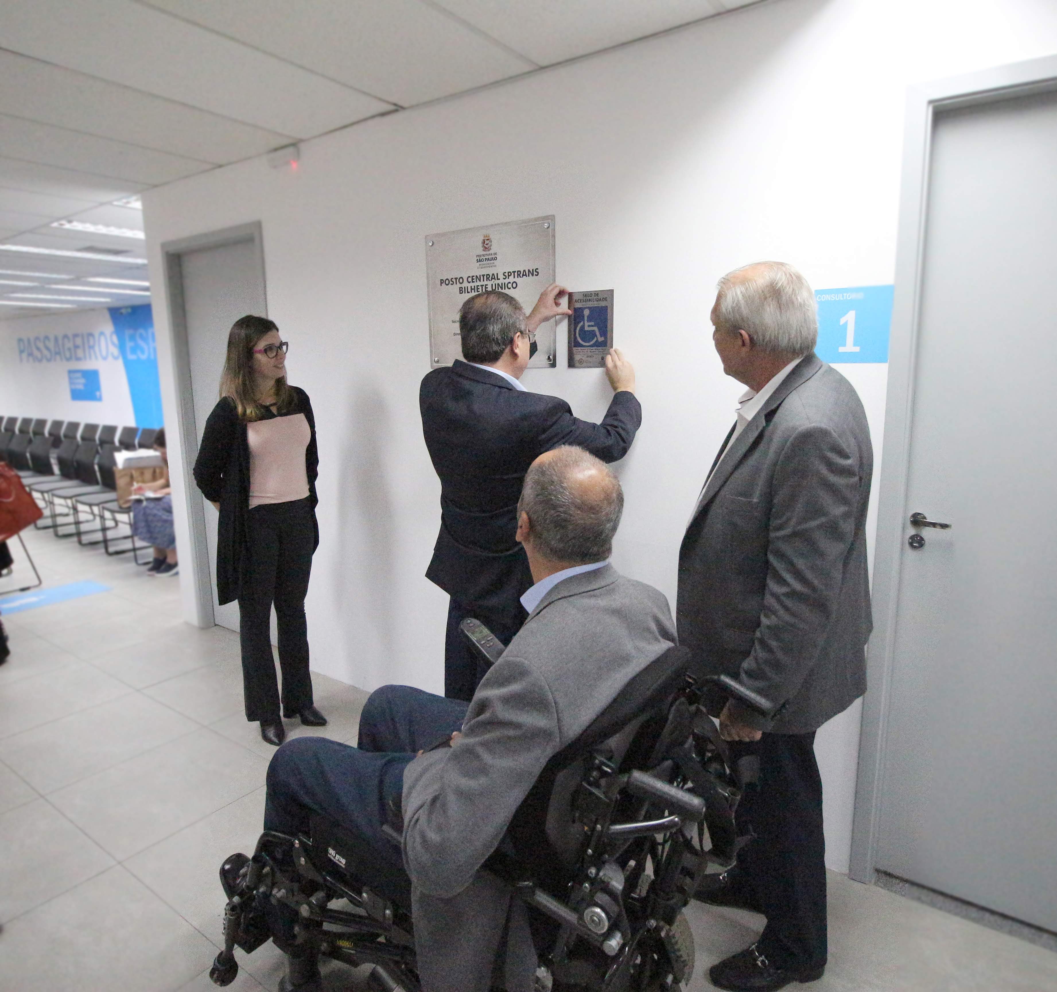 Quatro pessoas, uma delas colocando uma placa de selo de acessibilidade arquitetônica na parede enquanto os outros observam, entre eles, um homem em uma cadeira de rodas motorizada